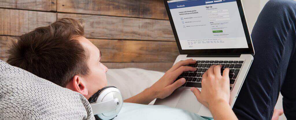 man laying on bed looking at facebook on laptop
