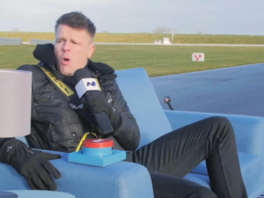 A man wearing a coat sitting on a blue sofa speaking into a microphone