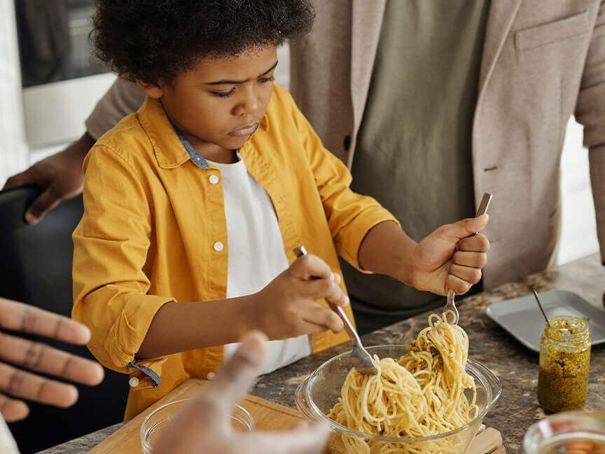a young boy wearing yellow is mixing spaghetti noodles in a bowl with forks