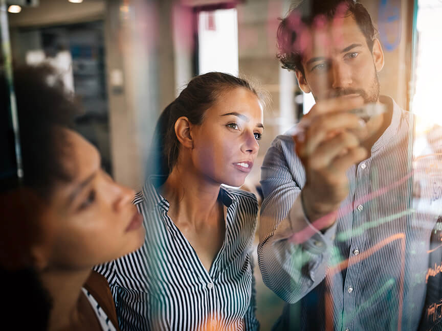 Two women and a man writing on a clear whiteboard