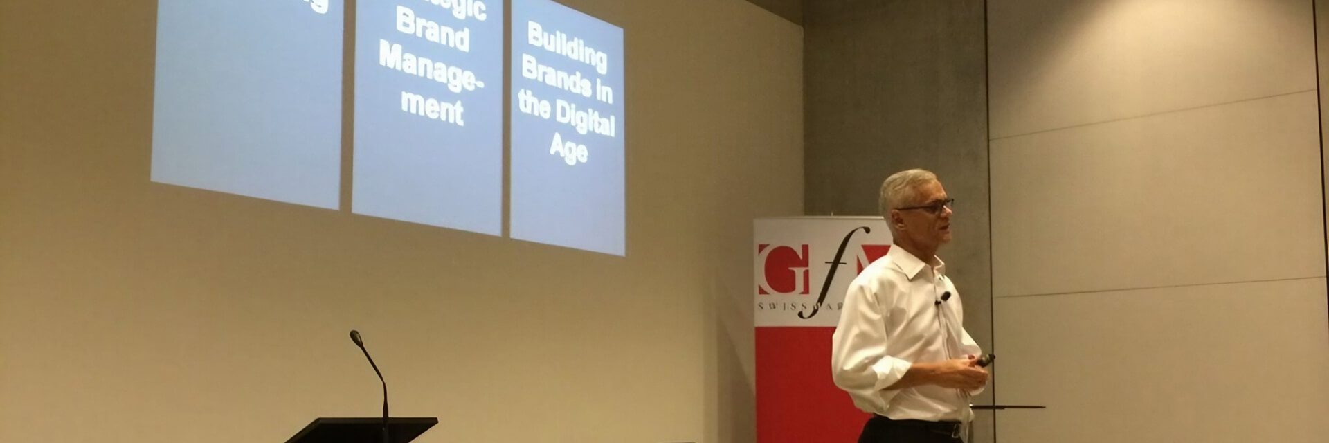 A man in a white shirt, black pants, and glasses presenting in front of a projected screen