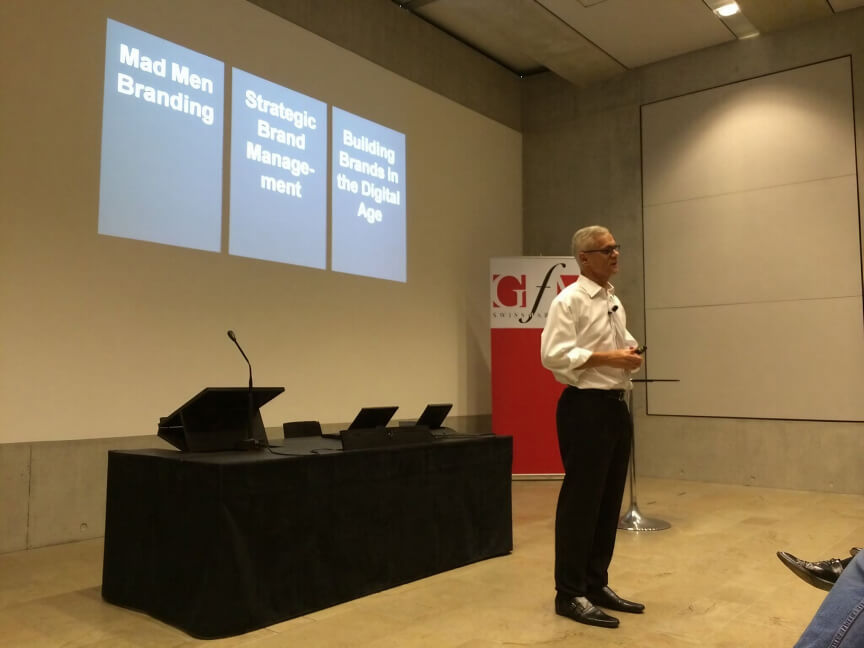 A man in a white shirt, black pants, and glasses presenting in front of a projected screen